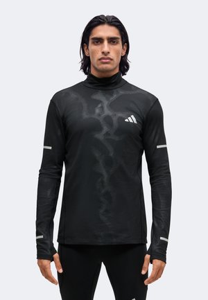 Mann trägt ein schwarzes, langärmeliges Sportoberteil mit hohem Kragen und feiner Musterstruktur, steht mit entspannten Armen.