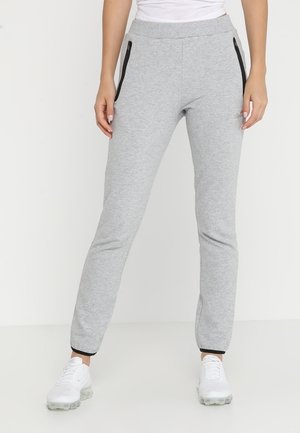 Kvinde iført lysegrå joggingbukser med sorte lynlåslommer, sort ankelsøm, hvide sneakers og en hvid top, stående mod en ensfarvet baggrund.