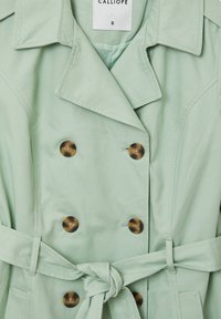 Trench coat verde menta con design doppiopetto, grandi bottoni marroni, vita cintrata e tessuto testurizzato. La fodera interna presenta delle strisce.