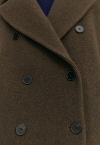 Bruine wollen jas met een dubbelzijdig ontwerp, voorzien van grote zwarte knopen en een gestructureerde afwerking vanuit een close-up perspectief.