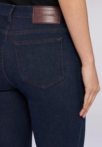 Vaqueros de mezclilla azul oscuro con un diseño ajustado, que presentan un bolsillo trasero rectangular y costuras rojas contrastantes. Etiqueta de cuero de Trussardi visible.