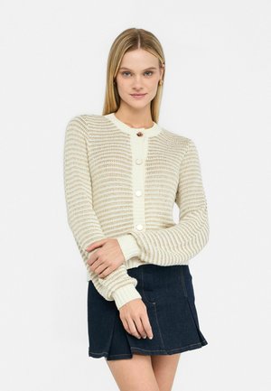 Cardigan in maglia di un beige chiaro e crema con righe orizzontali, chiusura con bottoni e polsini a costine. Abbinato a una gonna in denim scuro.