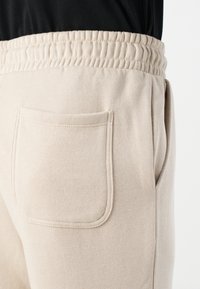 Beige sweatpants with elastic waistband, featuring a back pocket with stitched edges. The fabric appears soft with a smooth texture.