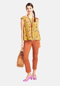 Gelber ärmelloser Oberteil mit Blumenmuster, kombiniert mit rostfarbenen, engen Hosen und pinken Schuhen mit Absatz. Eine braune Handtasche rundet das Outfit ab.