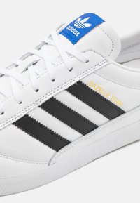 Hvit lær sneakers med svarte tre striper og "GAZELLE TEAM" i gull. Blå detalj på tungen, med hvite lisser og teksturert såle.