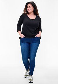 Black long-sleeve top, rounding neckline, paired with blue skinny jeans. Model wears athletic sneakers with green and white accents.
