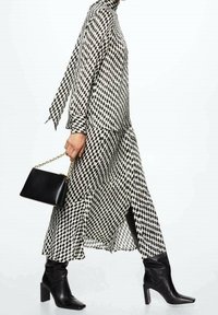 Schwarz-weißer kariert langer Kleid mit hohem Kragen, Seitenschlitz, langen Ärmeln und einer schwarzen Handtasche mit goldener Kettenriemen. Schwarze Knöchelstiefel.