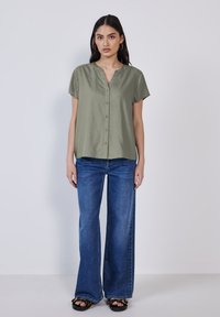 Blusa verde oliva a maniche corte con scollatura a V e bottoni frontali, abbinata a jeans in denim blu svasati e sandali neri.