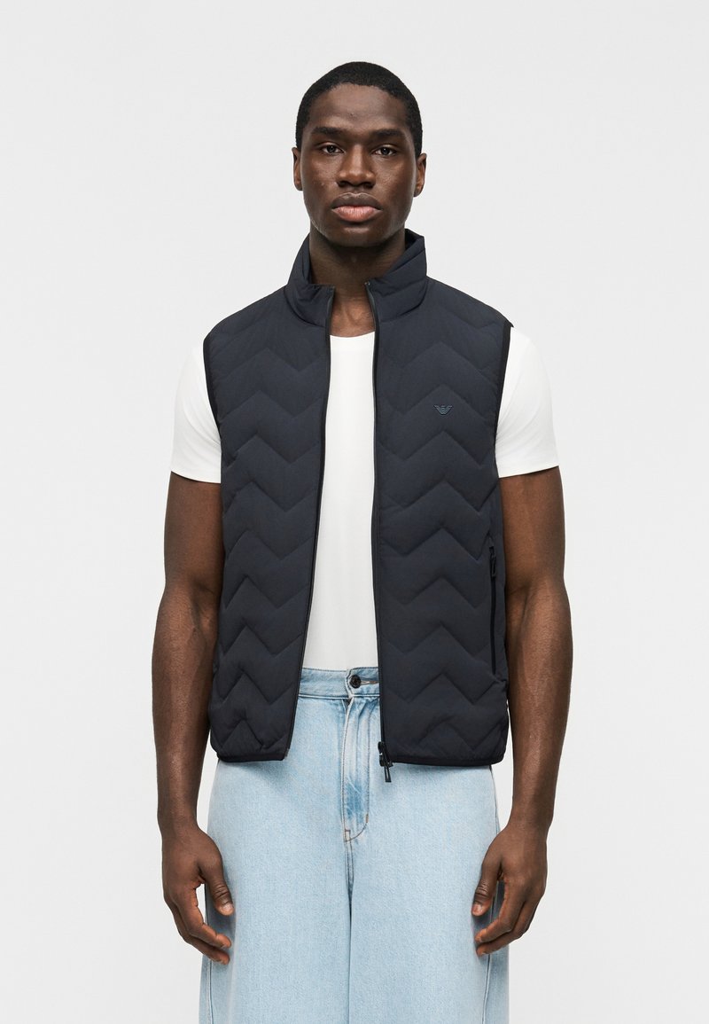 Άνδρας που φοράει μαύρο γιλέκο με ραφές πάνω από λευκό κοντομάνικο πουκάμισο και ανοιχτό μπλε τζιν, στέκεται μπροστά σε απλό λευκό φόντο.