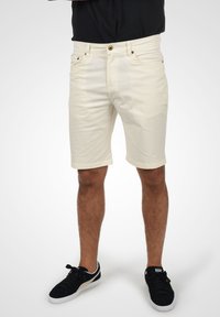 Lys beige bomullsshorts med rullekanter, fem lommer og gullknapplukking, brukt med svarte sneakers.