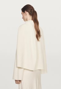 Cape en mélange de laine couleur crème avec manches larges, design drapé et texture lisse, portée par-dessus une robe assortie.
