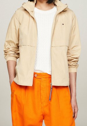 Person trägt eine beige Zip-up-Jacke über einem weißen strukturierten Hemd mit leuchtend orangenen Hosen, Hände in den Taschen.