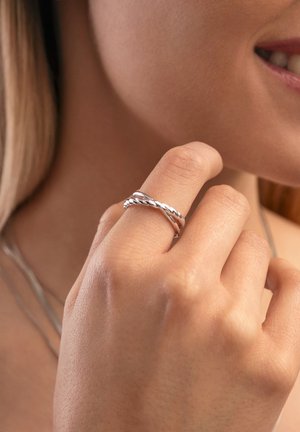 Primer plano de una mano cerca de un rostro sonriente mostrando un anillo de diseño plateado retorcido en el dedo índice.