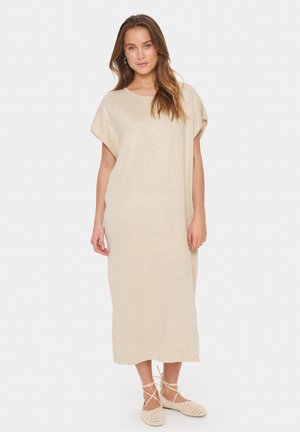 Beige knälång klänning med korta ärmar, rund halsringning, tillverkad av mjukt material; modellen bär ljusbeiga vävda skor.