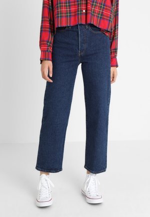 Persoon draagt donkerblauwe rechte spijkerbroek, witte hoge sneakers en een rood geruit overhemd met knopen en lange mouwen.