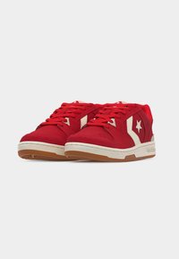 Paire de baskets rouges basses avec motif étoile et chevron blanc, semelles intermédiaires blanches et semelles extérieures couleur gomme, présentées en angle sur un fond uni.