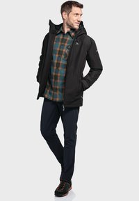 Zwarte waterdichte jas met capuchon, gedragen over een groen en bruin geruit shirt, gecombineerd met marineblauwe broek en zwarte wandelschoenen.