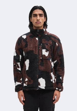 Uomo con capelli scuri che indossa una giacca in pile con zip e motivo nero, marrone, bianco e grigio, in piedi davanti a uno sfondo semplice.