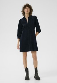 Modella in un vestito di velluto a coste blu scuro con zip frontale e maniche a sbuffo, che indossa stivali neri alla caviglia, in piedi davanti a uno sfondo bianco.