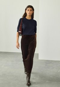 Donna che cammina all'interno, indossando una maglietta a maniche corte blu navy, una gonna in velluto marrone, stivali scuri e portando una borsa a tracolla rosso scuro.