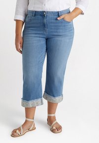 Jeans cortos de color azul claro con dobladillos deshilachados y acentos de patrón floral en los puños, combinados con sandalias planas beige.