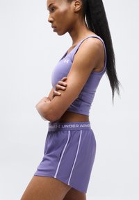 Γυναίκα με μακριά μαλλιά, φοράει μοβ αθλητικό τοπ και σορτς Under Armour που ταιριάζουν, με σταυρωμένα χέρια, κοιτάζοντας προς τα αριστερά σε απλό φόντο.