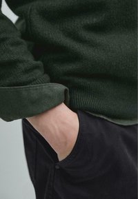 Tmavě zelený pletený svetr s ohrnutým rukávem, nošený přes černé kalhoty. Viditelná ruka spočívá v kapse, ukazující texturu a detaily designu.