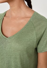 Woman wearing short-sleeve, green V-neck shirt, showing neck, collarbone, and part of braided hair.