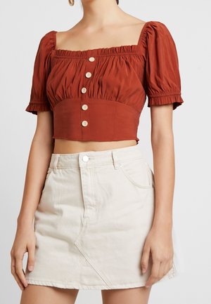 Kobieta w rdzawej, krótkiej bluzce z bufiastymi rękawami oraz beżowej spódnicy z wysokim stanem, z przeszyciami z przodu i kieszeniami.