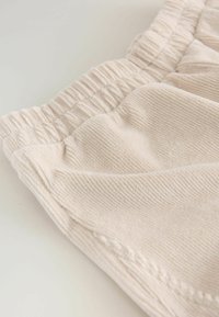 Material de culoare bej deschis, cu o textură ribbată, curea elastică și pliuri moi. Design simplu, vedere de aproape care pune în evidență calitatea materialului.