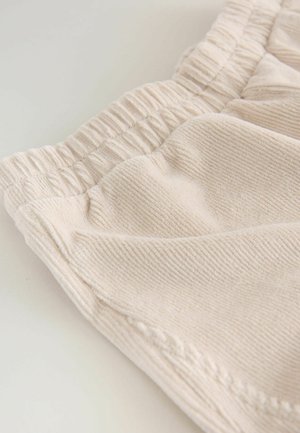 Lys beige stof med en ribbet tekstur, elastisk talje, og bløde folder. Enkel design, nærbillede der fremhæver materialekvaliteten.