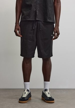 Man draagt een zwart overhemd met knopen en zwarte shorts met koord, witte sokken en zwarte sneakers, staand op een grijze vloer.