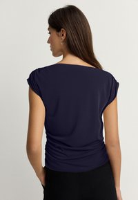 Tengerészkék rövid ujjú felső, összegyűrt vállakkal és laza szabással. Puha anyag, sima textúra, minimalista dizájn.