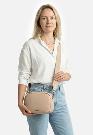 Sieviete baltā kreklā un gaiši zilās džinsās, turot bēša krāsas pleca somu ar platu siksnu uz vienkārša fona.