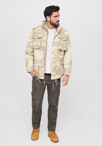 Beige Tarnjacke mit mehreren Taschen und Kordelzügen, kombiniert mit olivfarbenen Cargo-Hosen und hellbraunen Stiefeln. Darunter ein einfaches weißes T-Shirt.