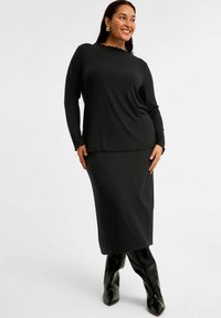 Musta ribbed pitkähihainen paita, jossa on kellotettu kaula-aukko, yhdistettynä yhteensopivaan maksihameeseen. Mallilla on päällään polvipituiset mustat kärkitossut.