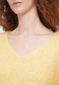 Κίτρινο πλεκτό πουλόβερ με λαιμόκοψη σε σχήμα V, που διαθέτει υφή με ανοιχτό πλεκτό σχέδιο. Κοντινή λήψη που τονίζει τη λαιμόκοψη και τις λεπτομέρειες του πλεξίματος.