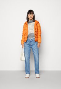 Casaco puffer em laranja brilhante, camisa de manga longa com padrão, jeans azuis, bolsa branca e sapatos brancos. Texturas suaves com um corte moderno.