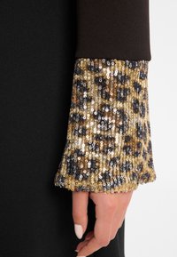 Schwarzes Kleid mit einem braunen Oberteil und einem glockigen Ärmel, verziert mit einem Leopardenmuster aus goldenen, braunen und schwarzen Pailletten.