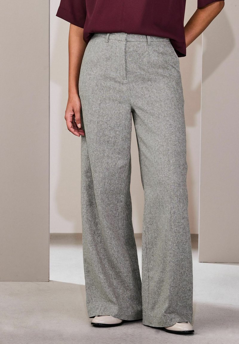 Mujer con pantalones de tiro alto y pierna ancha de color gris, combinados con zapatos de tono claro y una blusa de manga corta color granate oscuro.