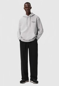 Ψηλός άνθρωπος που στέκεται με τα χέρια στις τσέπες, φορώντας μία ανοιχτού γκρι φούτερ από AllSaints, μαύρο παντελόνι και μαύρα αθλητικά παπούτσια με λευκές σόλες.