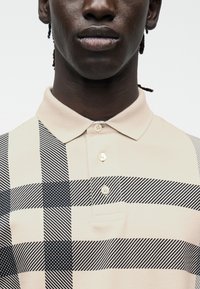 Gros plan d'une personne portant un polo à carreaux beige et noir avec trois boutons et un col, sur fond blanc.