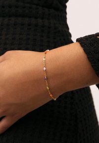 Pulsera de cadena de oro con cuentas multicolores en púrpura, naranja, amarillo y transparentes, llevada en una muñeca sobre un fondo de tela negra texturizada.