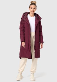Burgundy lång puffjacka med huva, med dragkedja fram, tryckknappar och quiltad design. Kombination med beige joggers och sneakers.