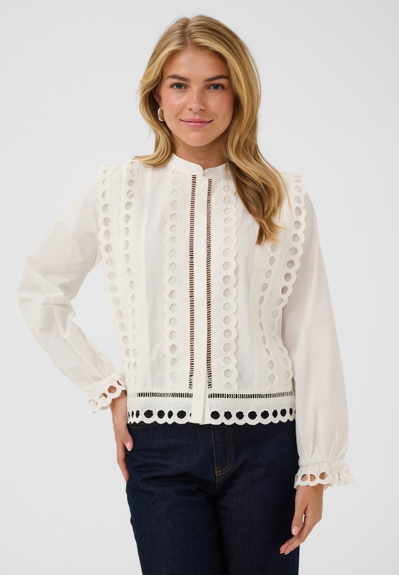 Femme blonde portant un chemisier blanc à manches longues avec des motifs en dentelle circulaires et un jean bleu foncé, debout avec une main sur la hanche.