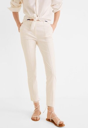 Femme portant un pantalon beige slim avec des coutures devant, une chemise crème nouée et des sandales plates blanches à brides, debout, les mains dans les poches.
