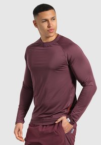Maglietta sportiva a maniche lunghe di un profondo bordeaux. Realizzata in tessuto liscio e elasticizzato con cuciture piatte e un design aderente. Presenta un logo discreto sul lato.