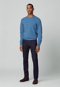 Jasnoniebieski sweter z okrągłym dekoltem wykonany z miękkiej dzianiny, zestawiony z ciemnymi dżinsami i brązowymi botkami za kostkę. Modelka stoi na tle jednolitego tła.