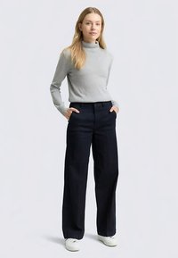 Pull à col roulé gris clair associé à un pantalon large bleu foncé. Le pantalon possède des poches et une légère brillance. Des baskets blanches complètent la tenue.