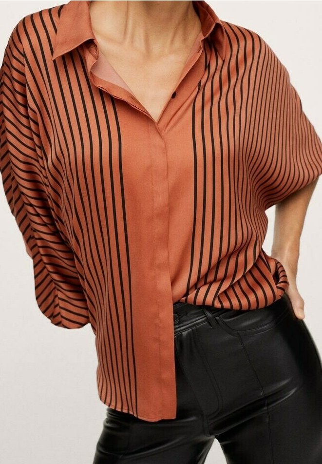 Blusa color ruggine con strisce verticali nere, chiusura frontale con bottoni e colletto. Design ampio e fluido con una texture morbida. Indossata con pantaloni neri.